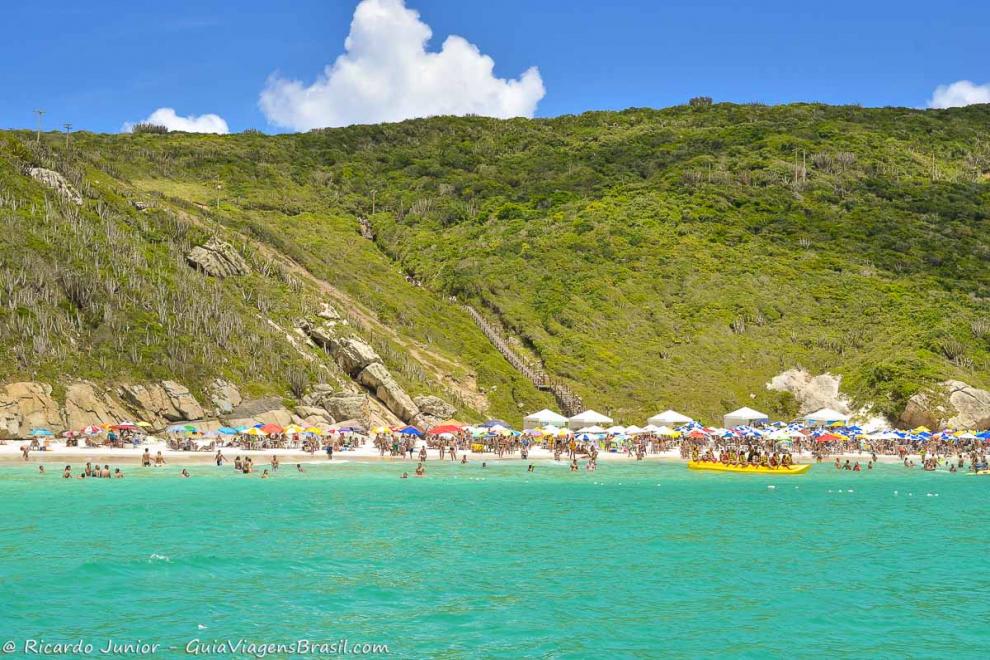 Imagem de um banana boat no mar e da praia repleta de turistas em Arraial do Cabo.